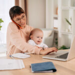 Junge Frau mit kleinem Baby auf dem Schoß im Vordergrund, sitzt am Schreibtisch vor einem Laptop, neben sich Dokumente und hinter sich Büromöbel; die junge Frau telefoniert und tippt gleichzeitig auf ihrem Laptop, das Baby lächelt.