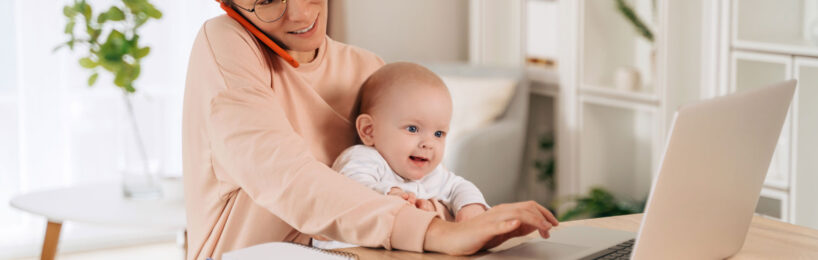 Junge Frau mit kleinem Baby auf dem Schoß im Vordergrund, sitzt am Schreibtisch vor einem Laptop, neben sich Dokumente und hinter sich Büromöbel; die junge Frau telefoniert und tippt gleichzeitig auf ihrem Laptop, das Baby lächelt.